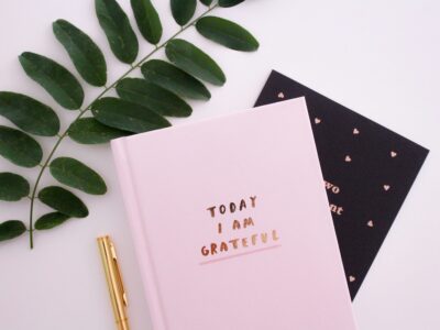 pink notebook, pen, and leaf on a light pink surface; notebook reads Today I Am Grateful