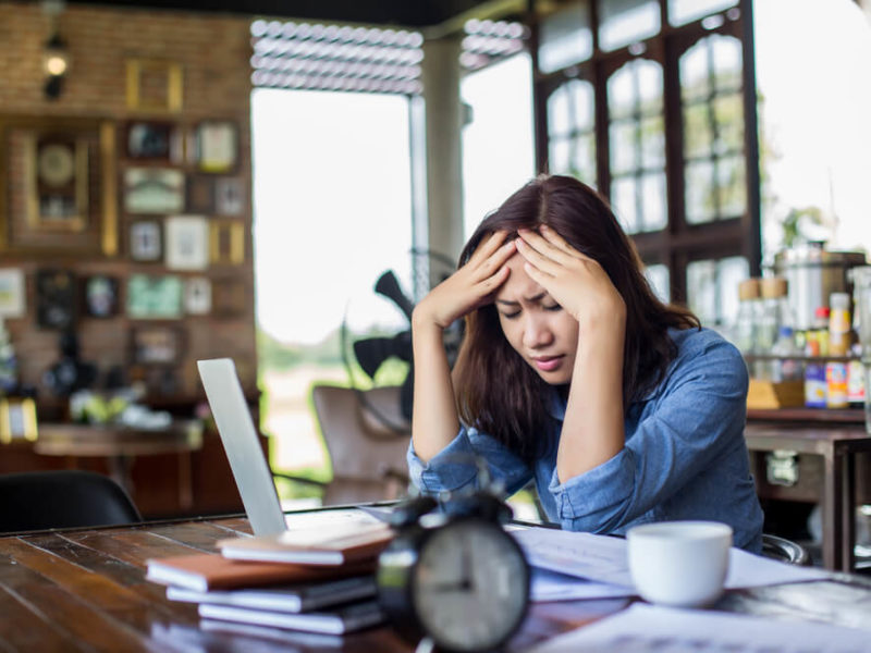 photo of woman sitting at desk with books and laptop, holding her head in pain