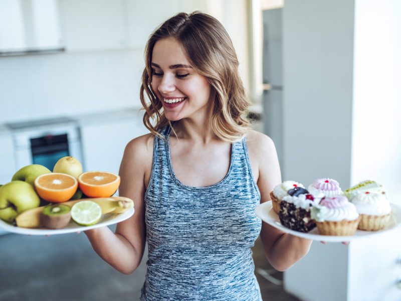 photo of a woman holding a plate of healthy food in one hand and junk food in the other