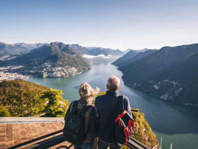 photo of mature couple wearing backpacks looking out at view of mountains and water