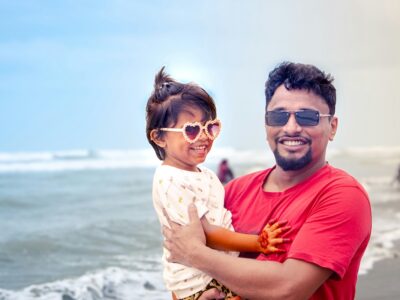 A joyful father and daughter share a heartwarming moment at the beach, standing near the gentle ocean waves under a bright blue sky.