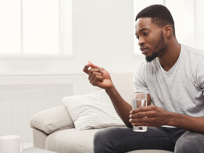 photo of man holding a glass of water in one hand and a pill in another, looking somber and contemplative