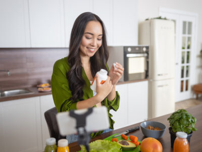 photo of a young woman standing in front of kitchen counter of healthy food looking at a supplement bottle