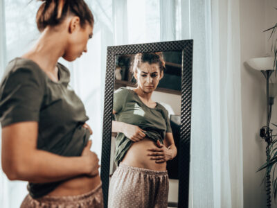 photo of woman holding bloated stomach, looking in mirror