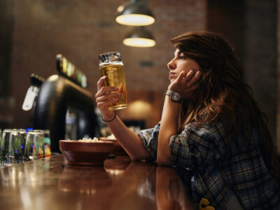 woman looking lonely at bar drinking a beer; alcohol and gut health concept