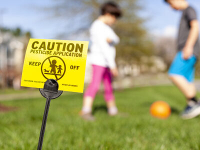 A yellow yard sign warning kids and pets of the recent pesticide spraying and advices them to stay away. Kids are playing soccer in the background regardless.