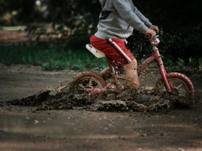 child in gray jacket and red pants riding red bicycle in mud puddle