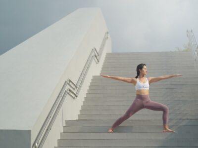 A woman practices yoga in a warrior pose on an outdoor staircase in a serene setting, emphasizing balance and strength, mindful movement