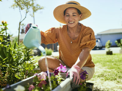 An elderly woman wearing a wide-brimmed hat is kneeling in her garden, watering her flowers with a watering can; benefits of gardening concept