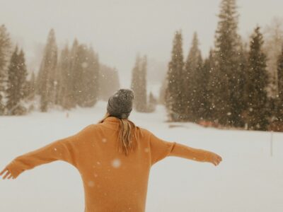 carefree woman standing in snow with arms outstretched and evergreen trees in the distance