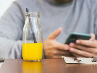 Man sitting at table with half drunk glass of juice, fork, knife, and napkin, no food, using his phone; juice cleanse, detox myths concept